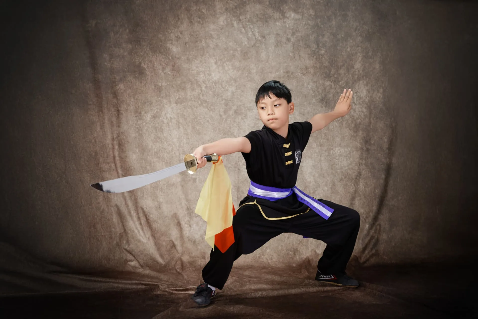 Student holding a broadsword in a low stance, wearing a purple sash